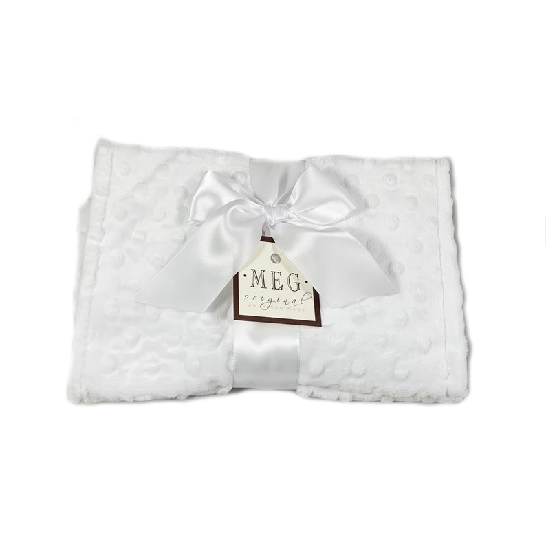 May include: A white baby blanket with a textured dot pattern, tied with a white satin ribbon and bow. A brown tag with the text "MEG original hand made" is attached to the ribbon.