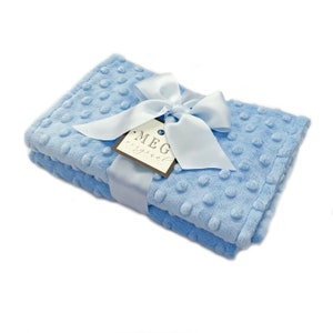 May include: A baby blue blanket with a textured, dotted surface. The blanket is folded and tied with a light blue satin ribbon and a white bow. A tag with the text "MEG original" is attached to the ribbon.