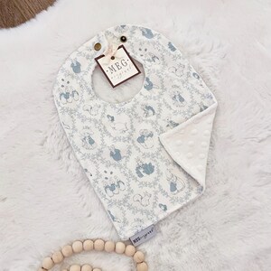 May include: A baby bib with a white and blue pattern featuring rabbits and squirrels. The bib has a rounded shape with a neck opening and a tag that reads "MEG original". The bib is made of soft fabric.