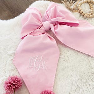 Könnte beinhalten: Eine große, hellrosa Schleife mit passendem Schal, bestickt mit weißen Initialen. Die Schleife ist in der Mitte gebunden und liegt auf einer weißen, flauschigen Oberfläche. Rosa Blumen sind unten.