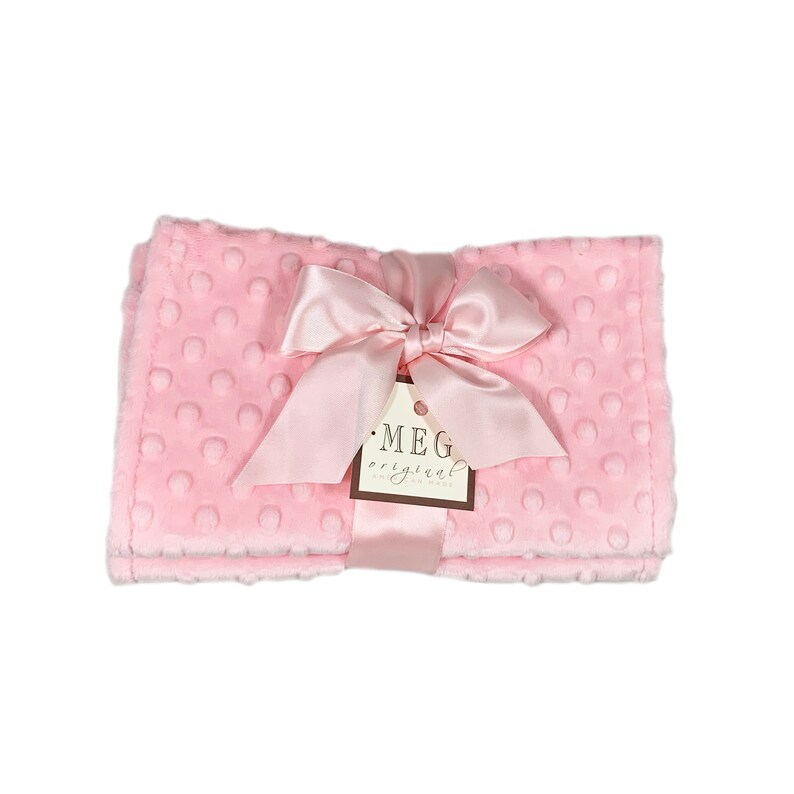 May include: A soft pink baby blanket with a textured dot pattern. The blanket is tied with a satin ribbon and a tag that reads "MEG original hand made". The blanket is folded and ready to be gifted.