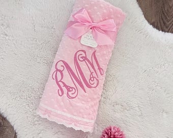 May include: A pink baby blanket with a textured, dotted surface and a white lace trim. The blanket is tied with a pink satin ribbon and features a monogram in a darker pink. The blanket is on a white fluffy rug.