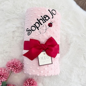 May include: Pink baby blanket with a white polka dot pattern and the name "Sophia Jo" embroidered in black. A red ladybug with a heart is embroidered above the name. A red bow is tied around the blanket and a brown tag with the text "MEG original" is attached.