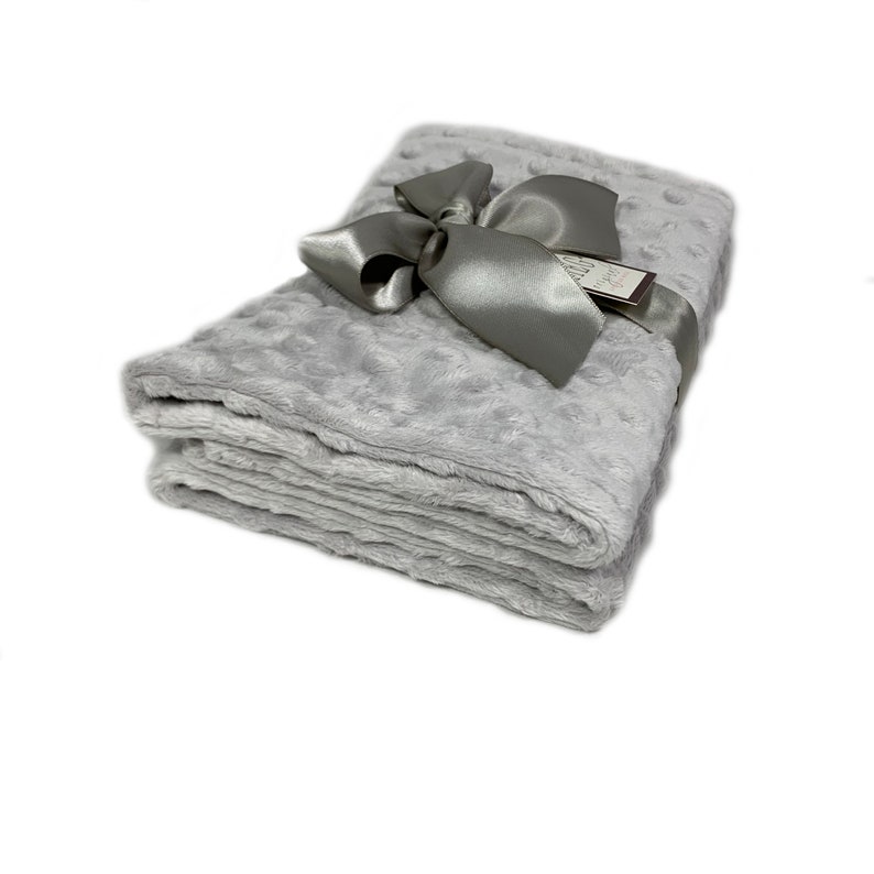 May include: A folded, light gray blanket with a textured surface and a satin ribbon tied in a bow. A small tag is attached to the ribbon. The blanket is made of a soft, plush material.
