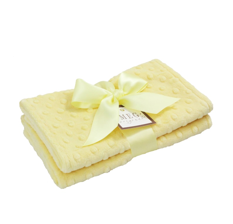 May include: A stack of two yellow blankets with a dot pattern, tied with a yellow satin ribbon and bow. A small tag with the text "MEG originals" is attached to the ribbon. The blankets are soft and fluffy.