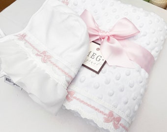 Puede incluir: Manta de bebé blanca con cinta de satén rosa y ribete de encaje blanco con lazos rosas. La manta está hecha de un tejido suave y texturizado. La manta se empaqueta con un gorro de bebé blanco con ribete de encaje rosa y lazos rosas.