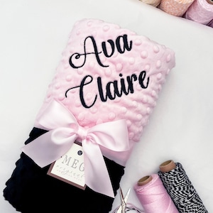 May include: A pink and black baby blanket with a pink satin bow. The blanket is personalized with the name "Ava Claire" in black embroidery. The blanket is made by MEG original.