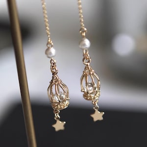 Puede incluir: Un par de pendientes de oro con una perla y un diseño de jaula. Cada pendiente tiene un pequeño encanto de estrella en la parte inferior.