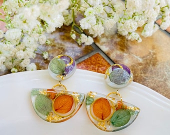 Könnte beinhalten: Ein Paar goldfarbene Ohrringe mit einem Halbkreisdesign. Die Ohrringe zeigen gepresste Blumen und Blätter in Grün-, Orange- und Lilatönen, die in klarem Harz eingeschlossen sind.