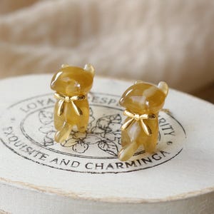 Peut inclure: Deux figurines de chat ambrées avec des nœuds dorés. Les chats sont posés sur une surface blanche avec l'inscription "Exquisite and Charming". L'arrière-plan est de couleur neutre et douce.