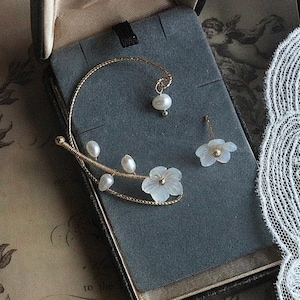 May include: A gold-toned ear cuff and stud earring set. The ear cuff features a floral design with pearls and a flower. The stud earring has a flower with a gold center. The jewelry is displayed in a gray box.