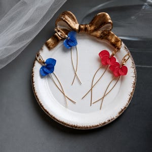 Op de afbeelding: Drie paar oorbellen met delicate, geconserveerde blauwe en rode hortensiabloemblaadjes bevestigd aan dunne, goudkleurige metalen draden. De oorbellen liggen op een rond, wit keramieken schaaltje met een decoratieve gouden strik aan de bovenkant.