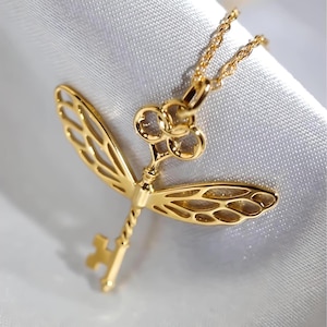 May include: A gold-toned necklace with a key pendant featuring a dragonfly design. The key has a twisted stem and a decorative flower-shaped top.