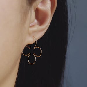May include: Close-up of a gold-coloured flower-shaped earring worn on an ear. The earring is a delicate, wire-frame design with four rounded petals. The background is a blurred, neutral colour.