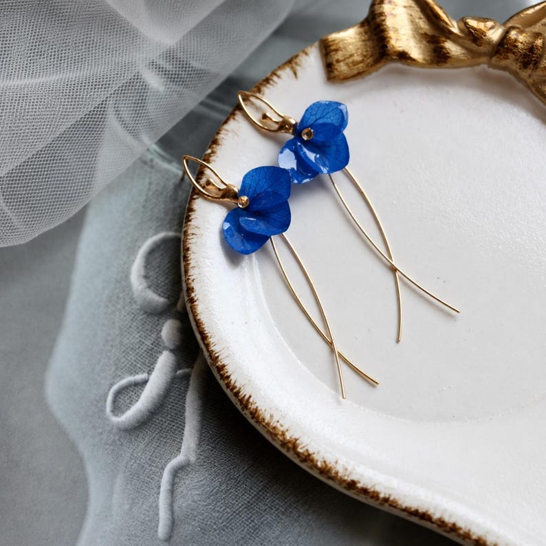 Op de afbeelding: Een paar oorbellen met felblauwe hortensiabloemblaadjes bevestigd aan dunne, gebogen goudkleurige metalen draden. De oorbellen liggen op een wit keramieken schaaltje met een gouden decoratief strikdetail aan de bovenrand.