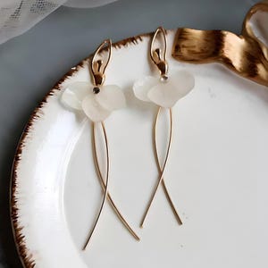 The white pressed flower earrings styled on a ceramic plate with a gold ribbon, creating a whimsical and romantic fairycore mood.