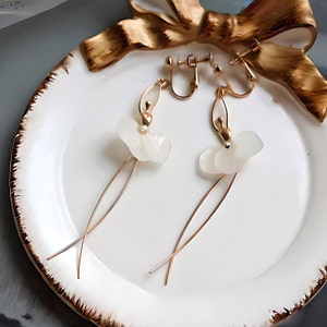 The clip-on version of the white Pressed Flower Ballerina Earrings, elegantly styled on a ceramic plate with a gold ribbon. Perfect for non-pierced ears.