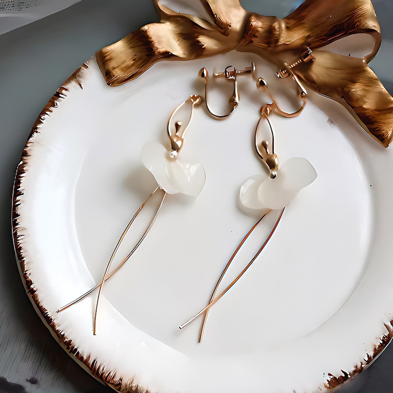 The clip-on version of the white Pressed Flower Ballerina Earrings, elegantly styled on a ceramic plate with a gold ribbon. Perfect for non-pierced ears.