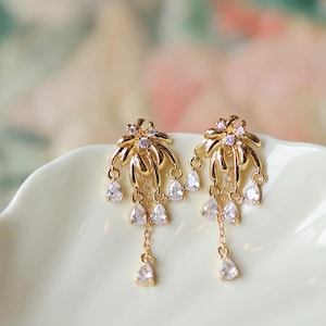 Puede incluir: Un par de pendientes dorados con un diseño floral. Cada pendiente presenta un grupo de pequeñas piedras claras en forma de lágrima colgando de delicadas cadenas. Los pendientes están colocados sobre un fondo suave y borroso.