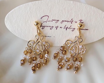 May include: Gold-toned chandelier earrings with dangling brown and clear beads. The earrings have an ornate design with small clear crystals. A card behind the earrings reads "Finding yourself is the beginning of a lifelong romance."