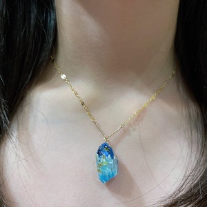 Peut inclure: Un collier en chaîne dorée avec un pendentif en résine bleu et blanc en forme de cristal. Le pendentif est parsemé de paillettes dorées.