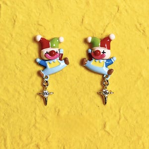 May include: A pair of whimsical clown earrings. Each earring features a colorful clown design with a red nose, a blue outfit, and a star-shaped charm. The earrings are set against a yellow background.