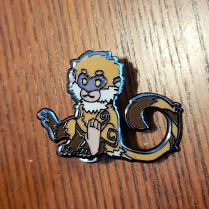 May include: A detailed enamel pin of a cartoon monkey in shades of tan, brown, and white, with a blue outline. The monkey is in a seated position with its hands resting on its lap. The pin has a glossy finish.