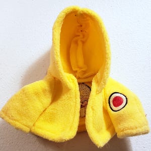 May include: A vibrant yellow hooded jacket, featuring a red and white circular emblem on the sleeve. The jacket is crafted from a soft, plush fabric and includes a hood. Designed for a doll or small toy.