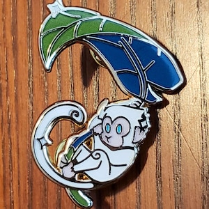 Könnte beinhalten: Emaille-Anstecknadel mit einem weißen Affen, der ein blaues und grünes Blatt hält. Der Affe hat blaue Augen und einen silbernen Umriss. Die Anstecknadel befindet sich auf einer Holzoberfläche.