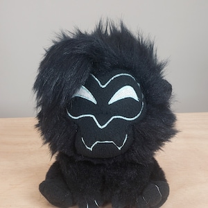 May include: Black plush toy with a white face mask design. The toy has a furry texture and is sitting on a wooden surface.