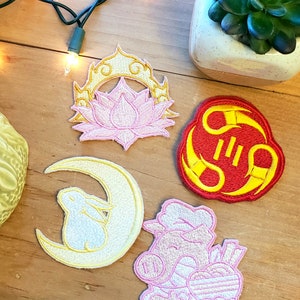 Puede incluir: Cuatro parches bordados para planchar con diferentes diseños. Una flor de loto rosa con un halo amarillo, un carácter chino rojo y amarillo, una luna creciente amarilla con un conejo blanco y un cerdo rosa que sostiene un corazón.