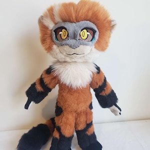 Puede incluir: Un peluche con forma de lémur con pelaje naranja y negro, una mancha blanca en el pecho y una máscara facial gris. La criatura presenta grandes ojos amarillos bordados, orejas puntiagudas y una cola larga y gruesa con la punta negra.