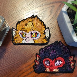 May include: Two embroidered patches of monkey faces. The first patch is yellow with orange fur and has blue eyes. The second patch is black with purple fur and has red eyes.