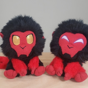May include: Two plush toys featuring black fur, red faces, and red limbs. The left plush has gold embroidered eyes, while the right plush has white embroidered eyes. Both have small red ears and a seated posture with visible red feet and hands.