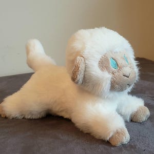 May include: A plush white yeti toy in a lying down position. The toy has a fluffy white body, a light brown face, ears, and paws, and turquoise embroidered eyes. The toy is on a gray surface.