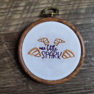 May include: A white embroidery hoop with a wooden frame and a purple and orange embroidered design of a bat with the text "one little spark".