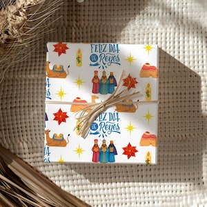 May include: A wrapped gift box with a festive design. The white paper features illustrations of camels, stars, candles, and the text "Feliz Día de Reyes" in blue. The box is tied with natural twine.