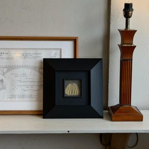 Op de afbeelding: Een kleine vierkante zwarte lijst met een gele pompoenillustratie met stippenpatroon, geplaatst naast een ingelijste architecturale tekening van een stenen brug en een hoge houten lamp met een heldere gloeilamp op een witte plank.