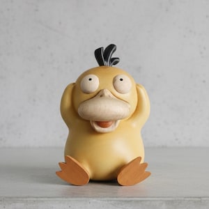 Op de afbeelding: Een gele Psyduck-beeldje met zijn handen op zijn oren. Het beeldje heeft een houten snavel en voeten, met zwart haar bovenop zijn hoofd. De ogen zijn groot en rond en de mond is open. Het beeldje staat op een grijze ondergrond.