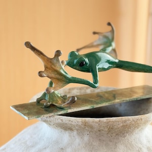 Può includere: Una scultura di rana verde con zampe palmate distese e occhi scuri, appollaiata su una barra di metallo piatta, rettangolare e screziata di verde e marrone, poggiata sul bordo di un vaso in ceramica bianco sporco e testurizzato.
