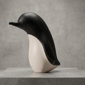 May include: A wooden penguin sculpture. The top is black, and the belly is cream-coloured. It stands on a grey surface against a neutral background. The sculpture has a smooth, curved design.