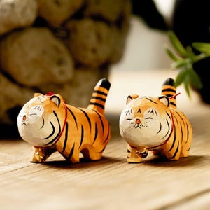 Può includere: Due statuette di tigre in legno con strisce arancioni e nere, ciascuna con un collare rosso e una piccola campana dorata. Le tigri hanno gli occhi chiusi, suggerendo uno stato di pace o sonno. Le statuette sono su una superficie di legno.