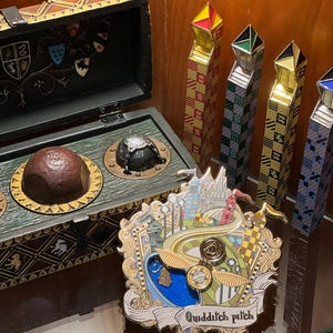Puede incluir: Un pin de metal dorado con una ilustración detallada de un campo de Quidditch, que presenta un snitch volador, un snitch dorado y el castillo de Hogwarts en el fondo. El pin tiene una pancarta blanca con el texto "Quidditch Pitch".