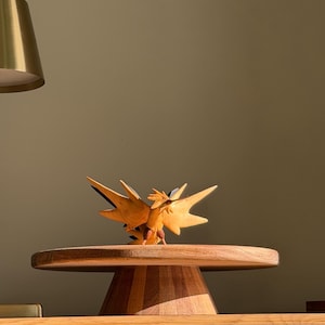 May include: A wooden bird sculpture with spread wings, displayed on a round wooden pedestal. The bird is light brown with darker details, and the pedestal has a wide, flat top. The sculpture is in a well-lit room.