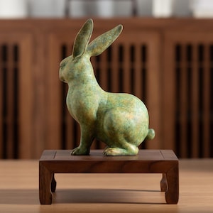 May include: A bronze-colored rabbit sculpture with long ears and a small tail, sitting on a small, dark wooden stand. The rabbit has a textured, mottled green and gold finish, and is posed in a seated position.
