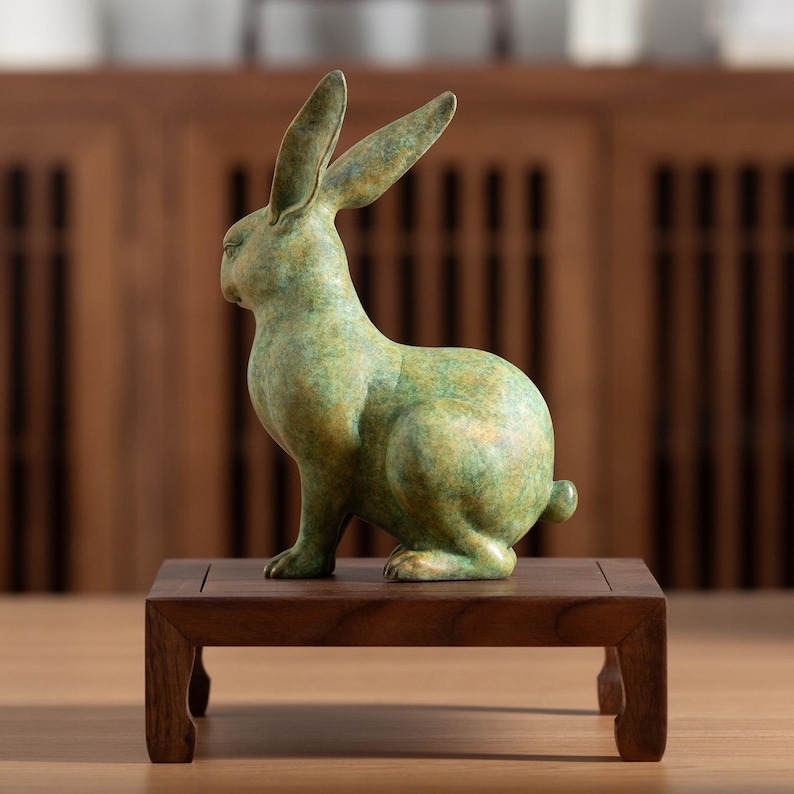 May include: A bronze-colored rabbit sculpture with long ears and a small tail, sitting on a small, dark wooden stand. The rabbit has a textured, mottled green and gold finish, and is posed in a seated position.