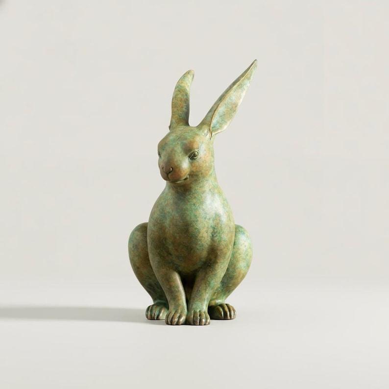 May include: A bronze-colored rabbit sculpture with a verdigris finish. The rabbit is depicted in a seated position with its ears perked up. The sculpture has a detailed texture and is set against a neutral background.