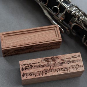 May include: Two wooden boxes with musical notation and text. One box is open, revealing a recessed interior. The other box displays the text "Concertino" and musical notes. A black clarinet is partially visible in the background.