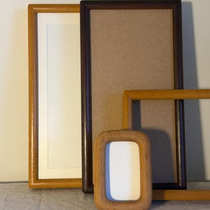 May include: A collection of rectangular picture frames in various wood tones. One frame is light oak with a white mount, another is dark brown wood, and a third is a smaller, rounded light wood frame with a white centre insert.