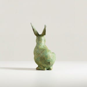 May include: A bronze-colored rabbit sculpture, viewed from the back, with long, upright ears. The sculpture has a textured surface and a slight green patina, suggesting age or weathering. The rabbit is in a seated position.